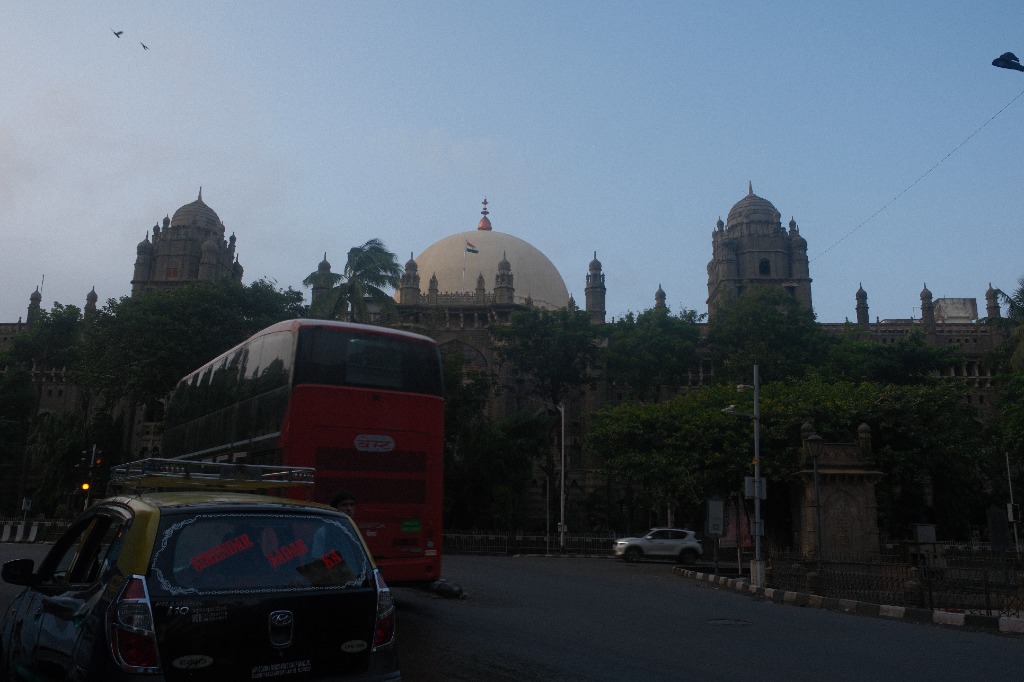 A red double-decker bus and a black-and-yellow taxi on a near-empty Mumbai street at first light, a domed heritage building behind them