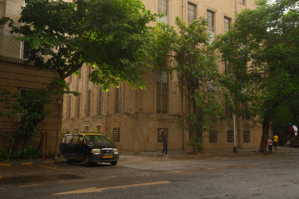 A black-and-yellow taxi parked on a rain-soaked street below a stone colonial facade screened by trees