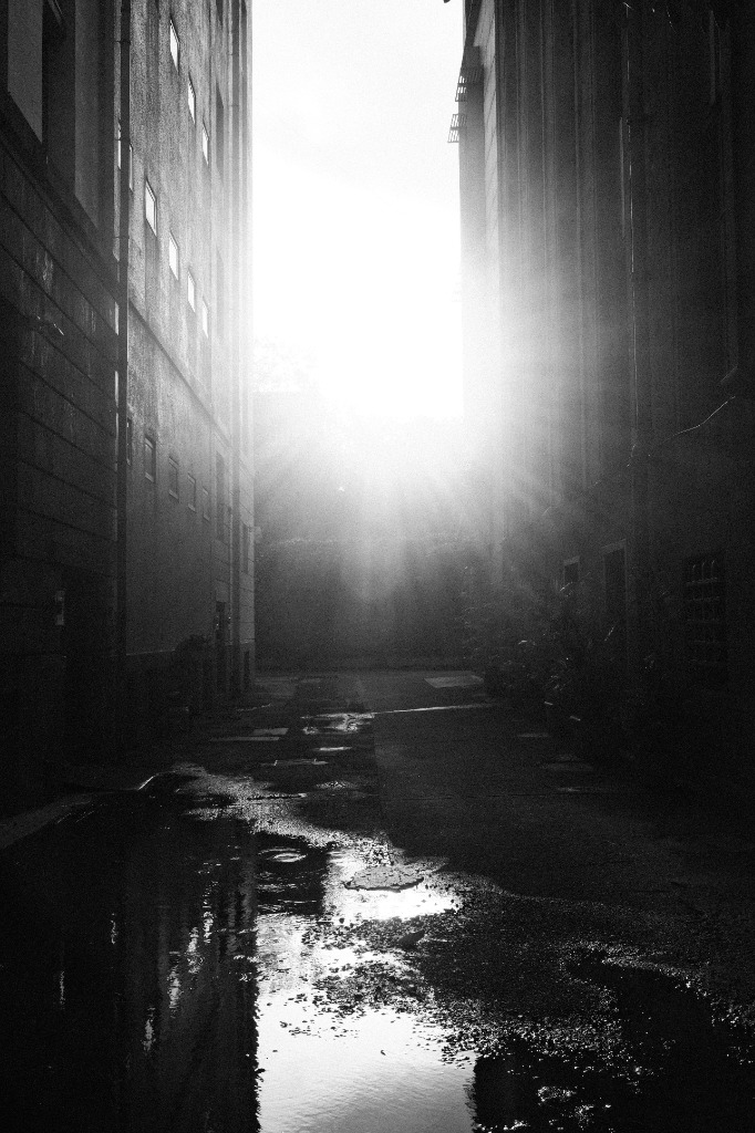 An alleyway with intense sunlight blooming and reflecting in a puddle.