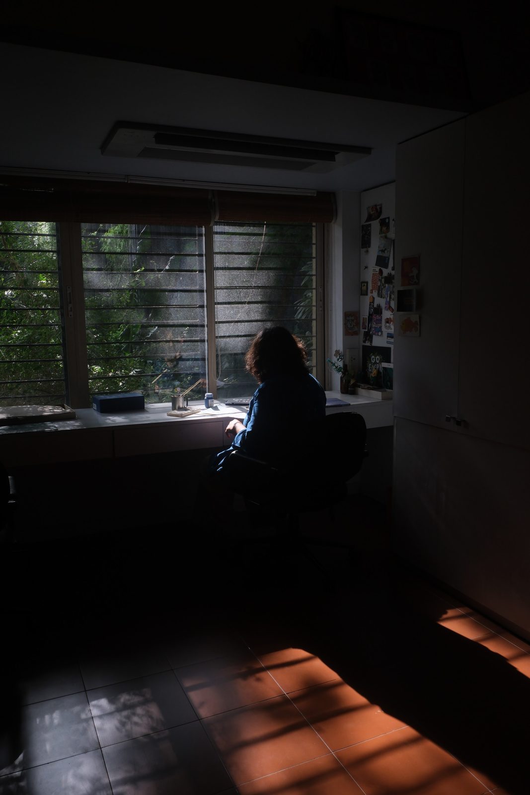 A woman sits at a desk running the length of a louvred window, silhouetted against the daylight, while a band of late sun falls across the tiled floor behind her