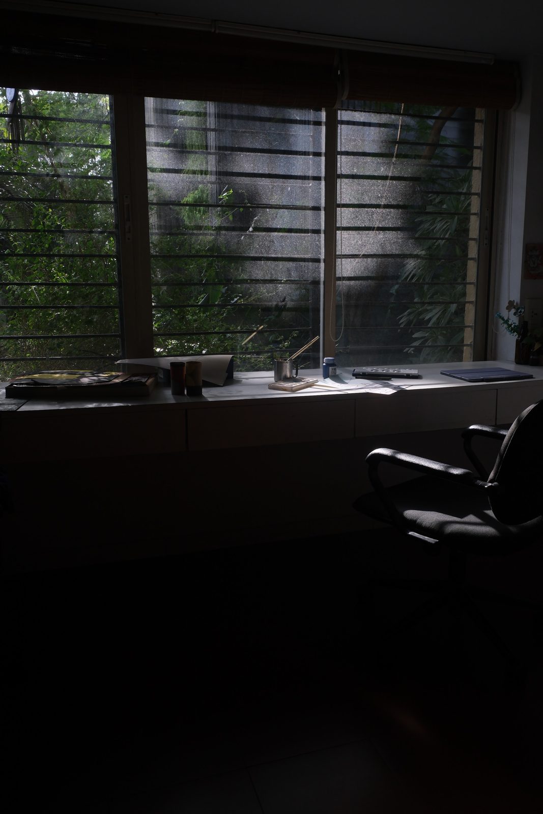 An empty room: a long desk under the window, an office chair pushed back, daylight coming through horizontal louvres with foliage beyond the glass